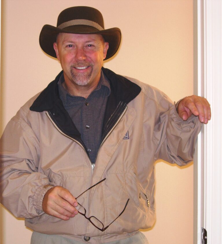 Smiling man in a beige jacket and cowboy hat indoors.