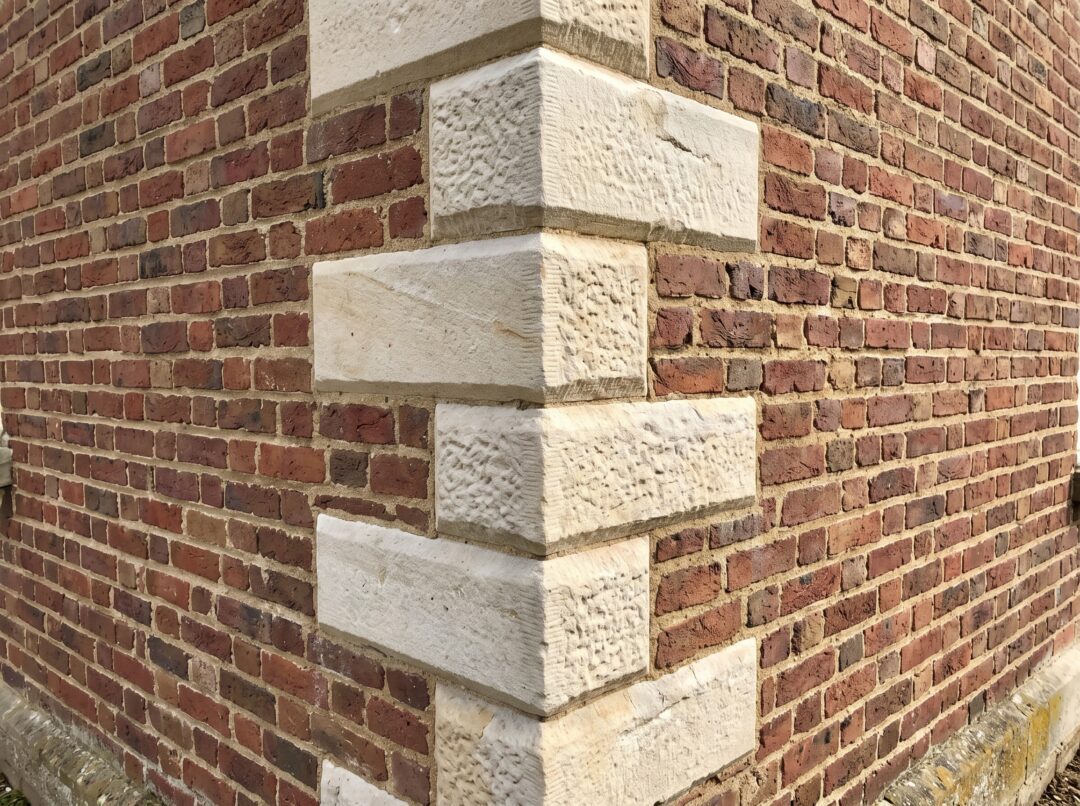 A close-up photograph of a traditional brick building's exterior corner, featuring vertically alternating, natural stone quoins interlocking with the surrounding weathered red brick masonry.